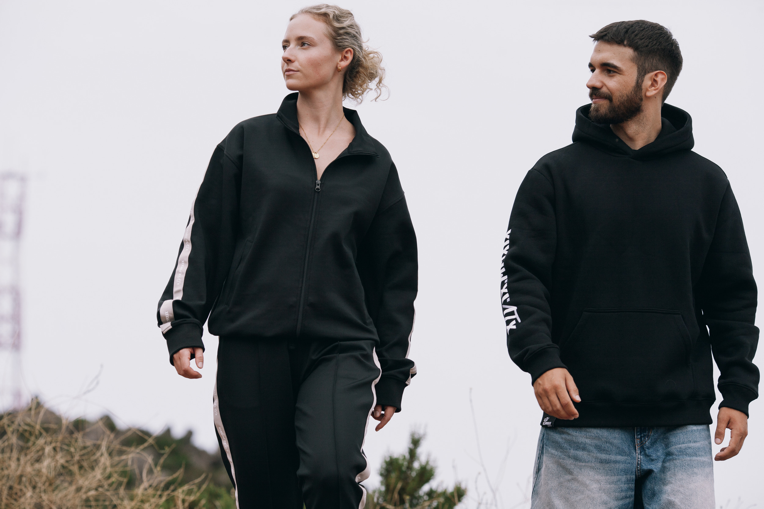 Two models walking toward the camera in a black track jacket and a black hoodie, hazy ridges behind them near Pico do Arieiro, Madeira