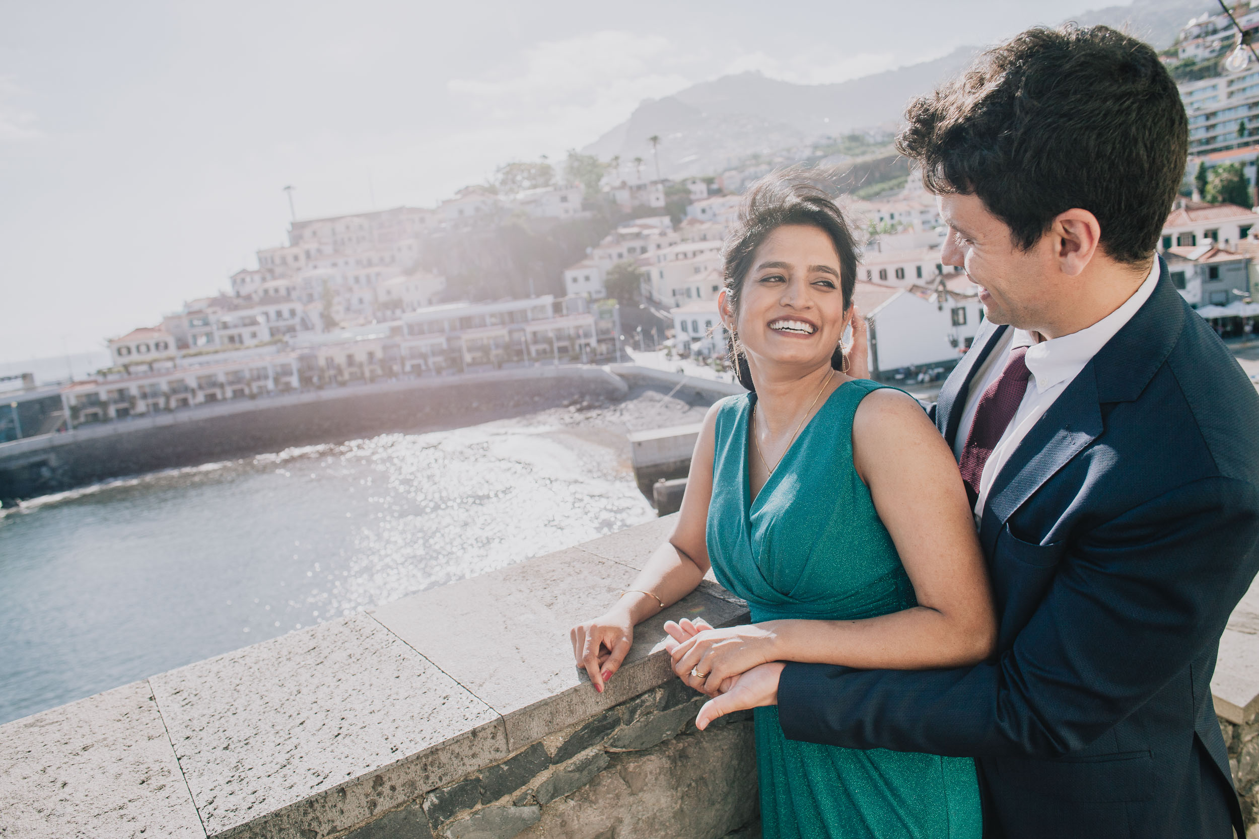 Couple on the sea wall above Câmara de Lobos bay, she is laughing in a green dress, he is in a navy suit, Madeira