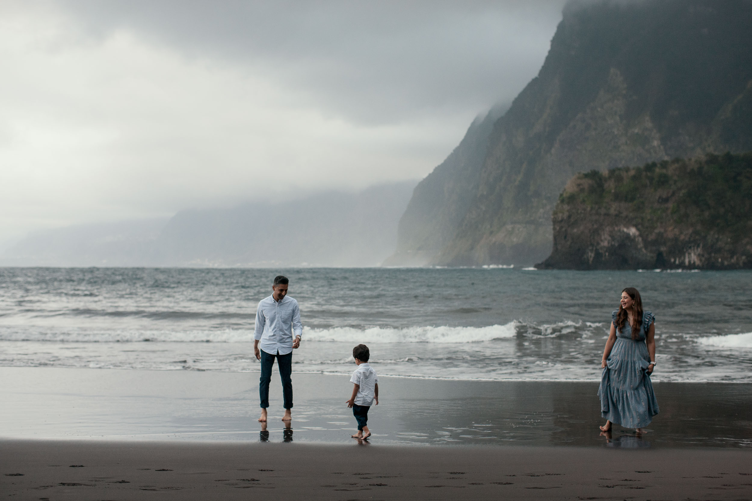 Family walking barefoot on a black sand beach on Madeira, cliffs in the mist behind them